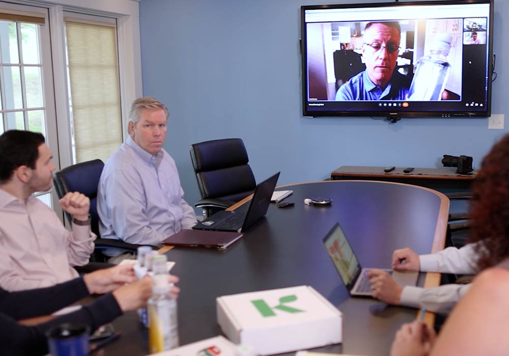 Employees in the office speak with a colleague on the screen who is working remotely about a marketing strategy.