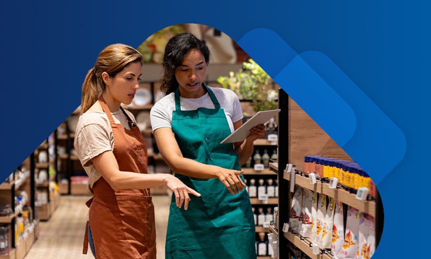 Two women wearing aprons collaborate in a grocery store aisle, one holding a tablet while pointing at the shelves stocked with products