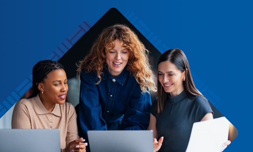 Three smiling women collaborate around a laptop against a blue background