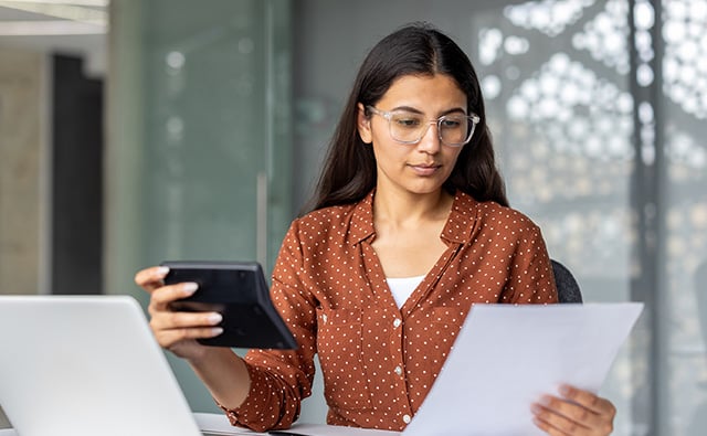 A focused business owner with glasses reviews documents and holds a tablet while seated at a desk in a professional setting