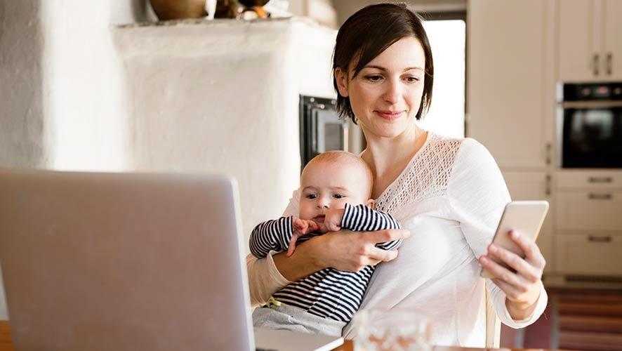 Una madre controla su cuenta de jubilación desde un dispositivo móvil mientras sostiene a su bebé.