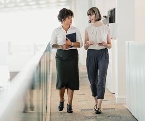 two women walking in a professional office setting carrying mobile phone and tablet