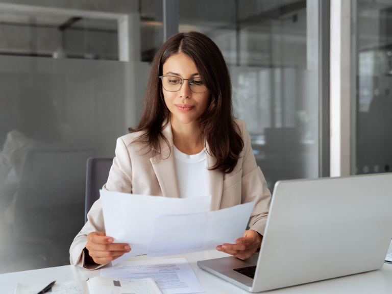Person seated at a desk in a glass-walled office reviewing printed documents beside an open laptop.