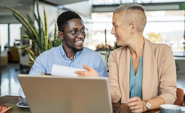 Two coworkers sit at a table with a laptop, reviewing documents and discussing work in a bright office setting.