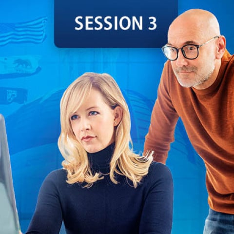 women sits at a desk looking at a computer monitors while a man stands beside her, leaning forward.