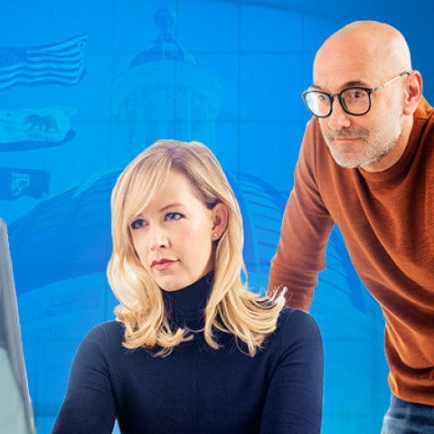 Two coworkers review a computer screen: a women seated at a desk and a mean leaning over her shoulder, both focused.