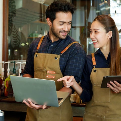 Two cafe employees wearing aprons standing behind a coffee counter, smiling while looking at a laptop and a tablet together in a warmly cafe setting