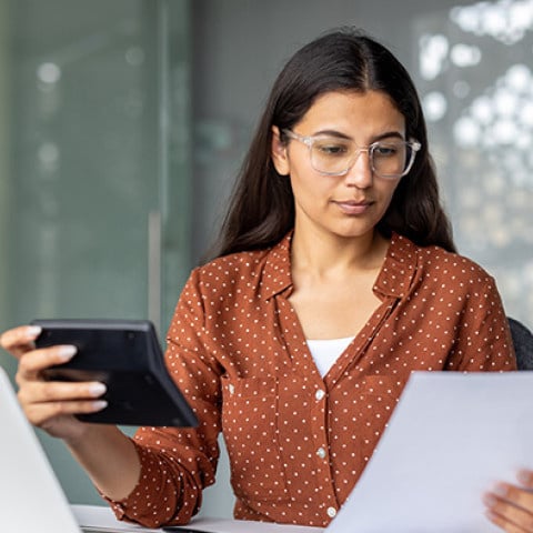 A focused business owner with glasses reviews documents and holds a tablet while seated at a desk in a professional setting