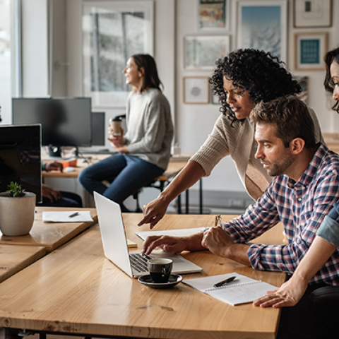 Colleagues in a modern office collaborates around a laptop, with one person pointing at the screen while others engage attentively.