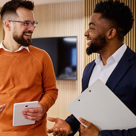 Two coworkers stand in an office setting discussing documents, with one holding a tablet and the other holding printed papers.