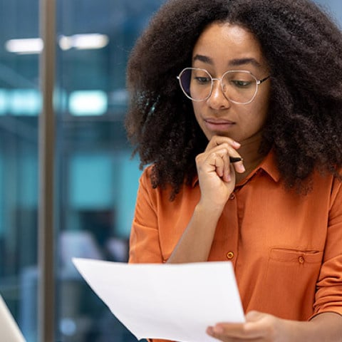 A person sits at a desk in an office setting, reviewing a printed document beside an open laptop.