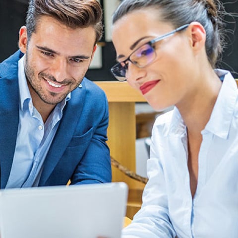 Two people sit together at a table reviewing information on a laptop in a modern workspace.