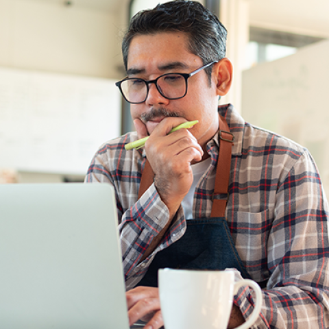  A person wearing a plaid shirt and apron sits at a desk working on a laptop, with a coffee mug in the foreground and a bright office environment in the background.