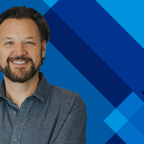 A person with a beard wearing a blue button-up shirt stands against a blue geometric background with diagonal shapes, facing the camera with arms crossed.