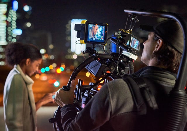 A camera operator films a person in an urban setting at night, with professional video equipment and city lights visible in the background.