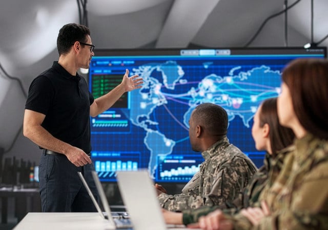 A presenter gestures toward a large digital world map and network monitoring dashboard while a group of uniformed personnel and colleagues watch from a conference table with laptops.