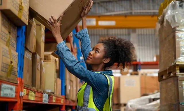 A female warehouse worker handles a box while storing items on shelves.