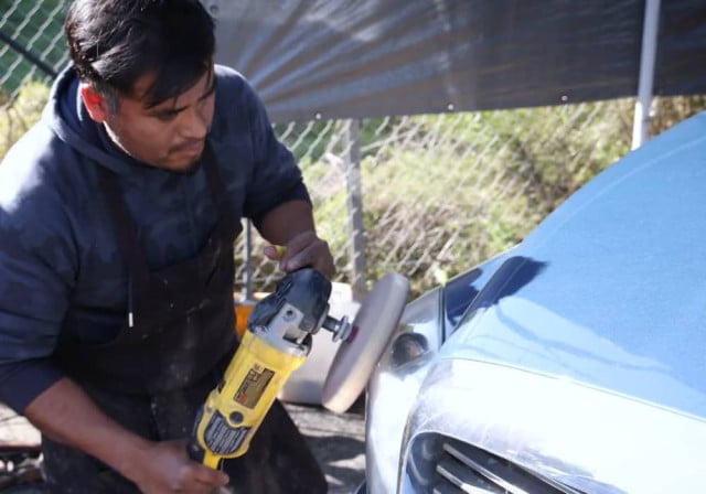 dark haired man in blue uniform uses buffing equipment on automobile