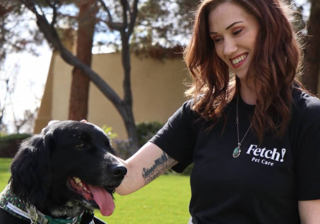 Mujer joven de cabello oscuro que está usando una camiseta de “FETCH! Pet Care” acaricia a un perro negro