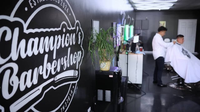 logotipo de champions barbershop en la pared izquierda, barbero lejano y cliente masculino en silla cortándose el pelo