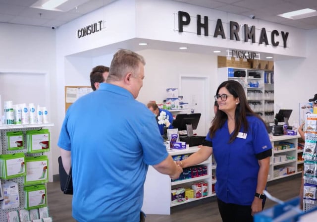 dark-haired woman in blue shirt shakes hands with older man with greying hair outside a pharmacy counter
