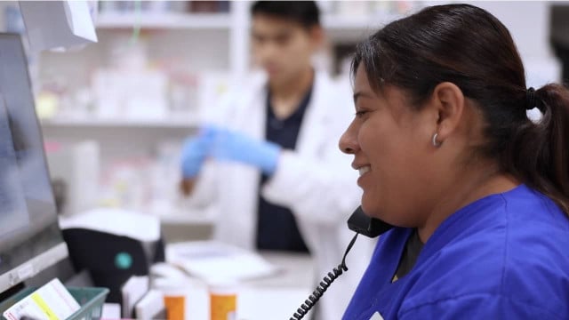 dark haired woman in blue pharmacy scrubs has phone pressed to ear while working on computer screen, close up