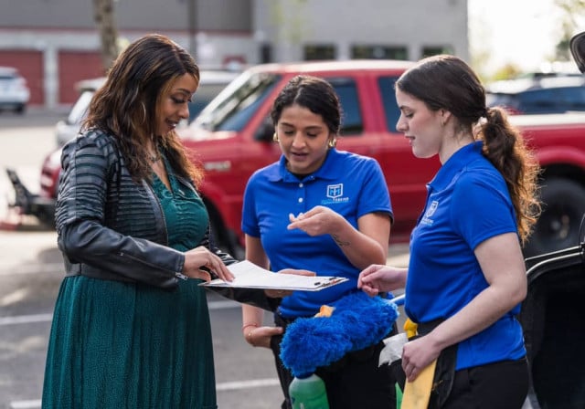 dos jóvenes trabajadoras de limpieza con camisas azules observan un portapapeles con notas que sostiene una mujer afroamericana con un vestido largo verde