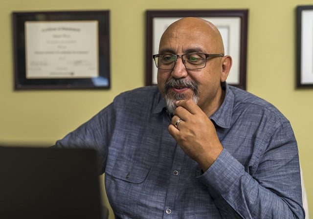 bearded man with glasses in a blue shirt rests his chin on his hand as he looks at a computer