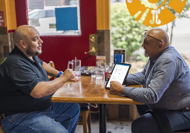 older man seated at a table in a restaurant with vibrant colored walls shows business info on an ipad to a somewhat younger man. They appear to be having a business meeting. 