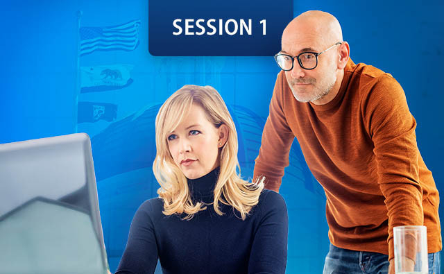women sits at a desk looking at a computer monitors while a man stands beside her, leaning forward.