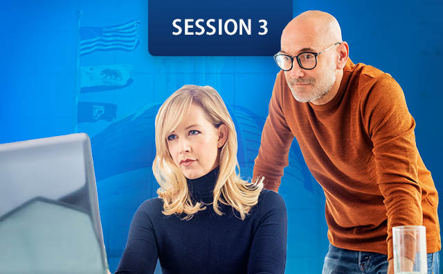 women sits at a desk looking at a computer monitors while a man stands beside her, leaning forward.