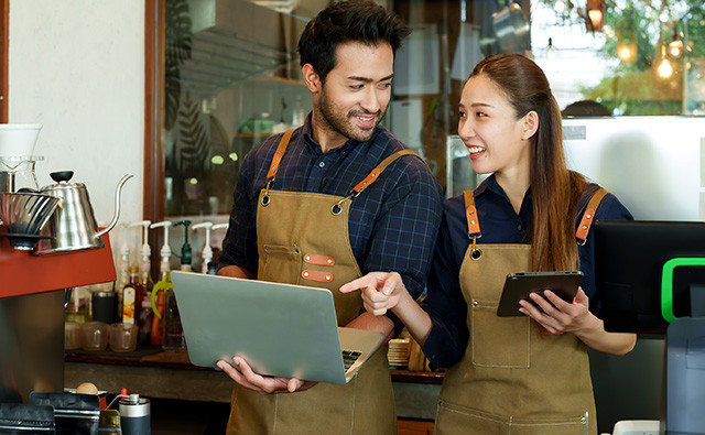 Two cafe employees wearing aprons standing behind a coffee counter, smiling while looking at a laptop and a tablet together in a warmly cafe setting