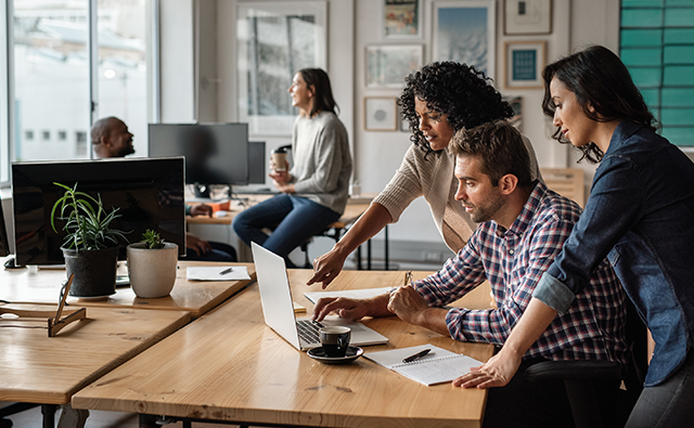 Colleagues in a modern office collaborates around a laptop, with one person pointing at the screen while others engage attentively.