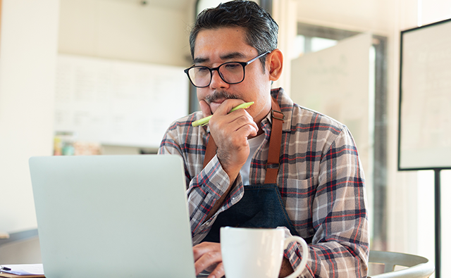  A person wearing a plaid shirt and apron sits at a desk working on a laptop, with a coffee mug in the foreground and a bright office environment in the background.
