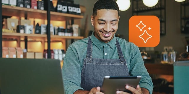 Café worker wearing an apron uses a tablet at the counter, with shelves of products and equipment visible in the background.
