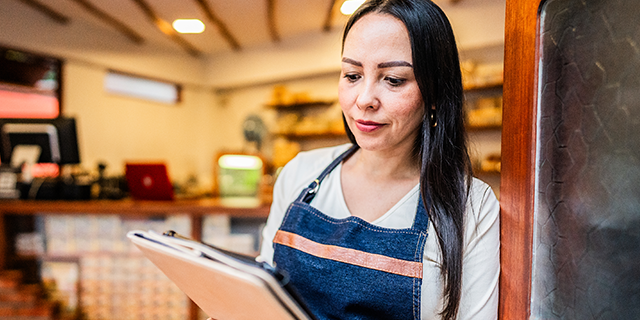 Person in a café wearing an apron reviews a clipboard order sheet at the counter with shelves and equipment in the background.