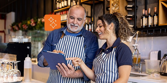 Two café workers wearing striped aprons stand at a counter reviewing a tablet together, with glassware, bottles, and coffee equipment visible in the background.