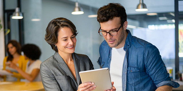 Two people in a modern office stand side by side, looking at and interacting with a tablet. One person wears a gray blazer, and the other wears glasses and a blue shirt. In the background, a softly blurred workspace shows additional people and hanging lights, suggesting a collaborative work environment.