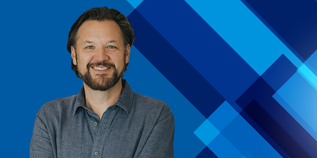 A person with a beard wearing a blue button-up shirt stands against a blue geometric background with diagonal shapes, facing the camera with arms crossed.