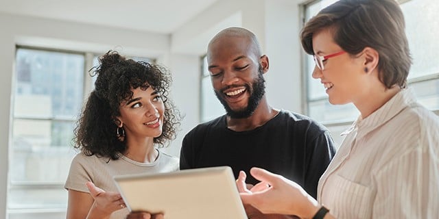 Three people stand together in a bright room, looking at and discussing content on a tablet.