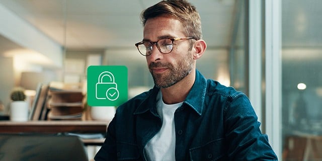 Person wearing glasses working on a laptop in an office setting, with a green padlock icon on the screen indicating security or data protection.
