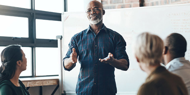 A person stands in front of a whiteboard, gesturing with both hands while speaking to three seated people in a bright room with large windows.