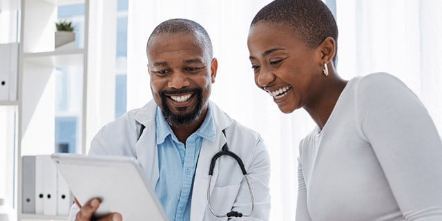 A healthcare professional wearing a white coat and stethoscope and another person stand together indoors, looking at a tablet in a bright, modern office setting.