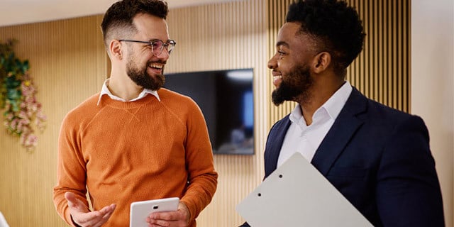 Two people stand indoors holding a tablet and a folder, facing each other in conversation in a modern office setting.