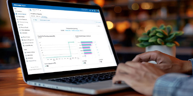 A laptop on a desk displays an analytics dashboard with charts and graphs as a person types on the keyboard in a softly lit workspace.