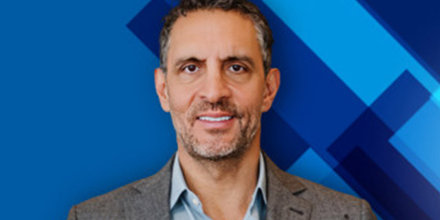 A person is shown from the shoulders up against a blue geometric background, wearing a blazer and collared shirt and facing the camera.