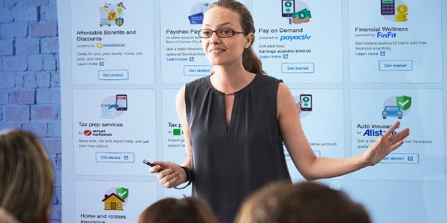 A presenter stands in front of a large screen displaying benefits and financial service options, gesturing while addressing an audience in a professional setting.