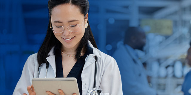 A healthcare professional wearing a white coat and stethoscope looks down at a tablet in a clinical or workplace setting with people and equipment softly visible in the background.