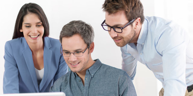 Three people lean toward a laptop screen together, collaborating in a bright workspace with a white background.