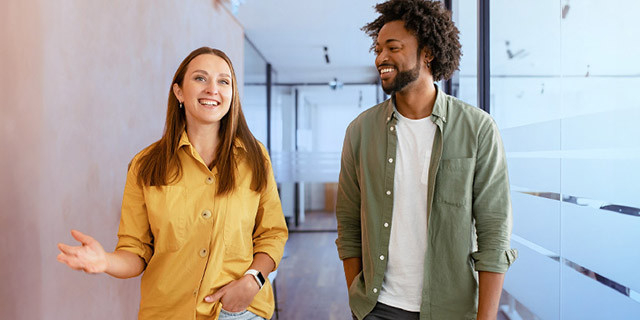 Two people walk and talk together down a bright office hallway, wearing casual work attire and engaging in conversation.